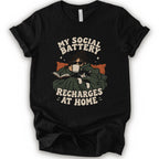 A black short-sleeve graphic t-shirt features a cozy illustration of a person wrapped in a green blanket reading a book with a black cat, surrounded by the text 'MY SOCIAL BATTERY RECHARGES AT HOME' in a beige retro font.