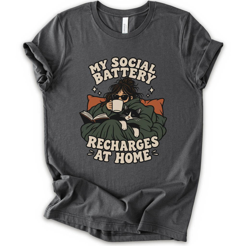 As asphalt short-sleeve graphic t-shirt features a cozy illustration of a person wrapped in a green blanket reading a book with a black cat, surrounded by the text 'MY SOCIAL BATTERY RECHARGES AT HOME' in a beige retro font.