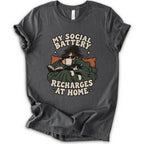 As asphalt short-sleeve graphic t-shirt features a cozy illustration of a person wrapped in a green blanket reading a book with a black cat, surrounded by the text 'MY SOCIAL BATTERY RECHARGES AT HOME' in a beige retro font.