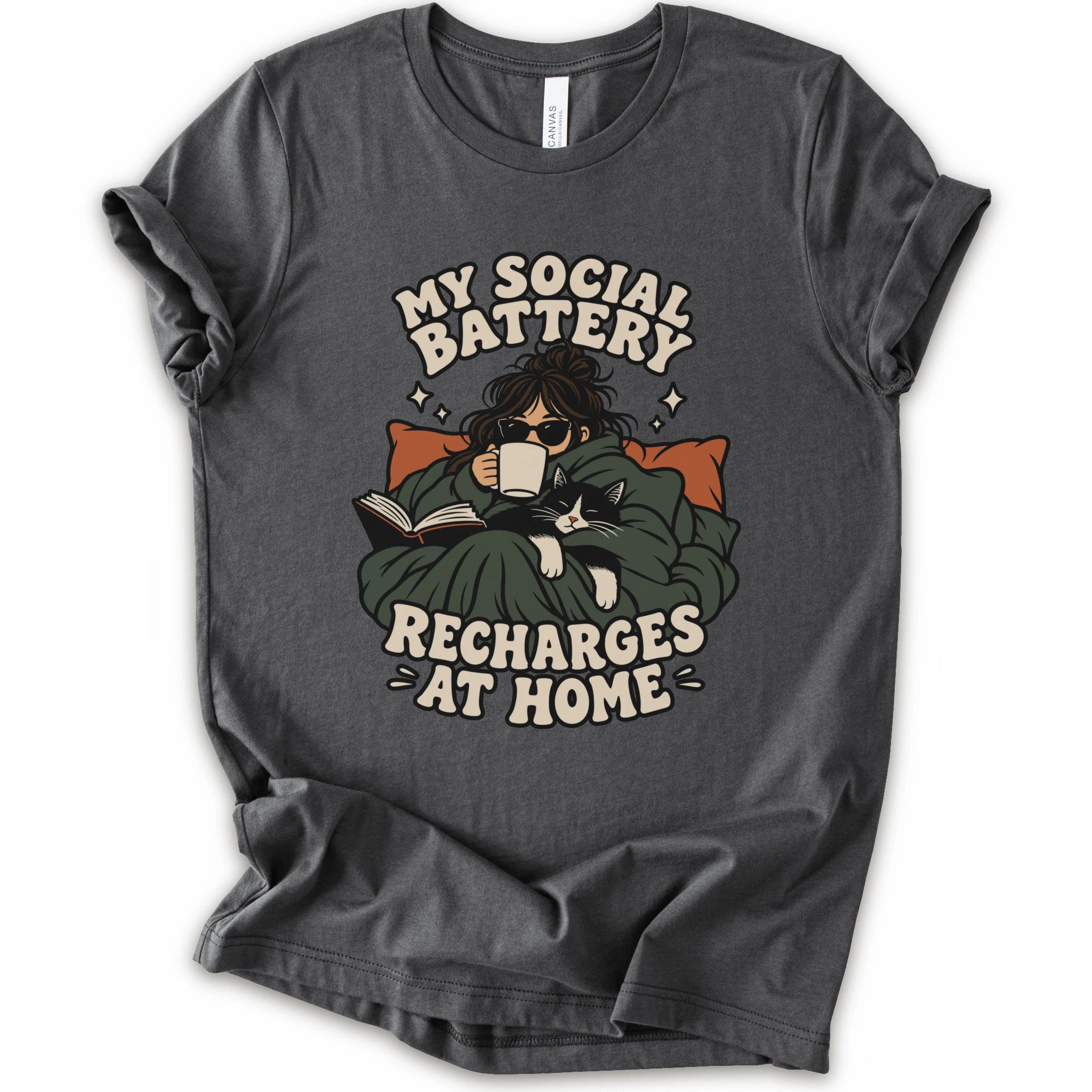As asphalt short-sleeve graphic t-shirt features a cozy illustration of a person wrapped in a green blanket reading a book with a black cat, surrounded by the text 'MY SOCIAL BATTERY RECHARGES AT HOME' in a beige retro font.