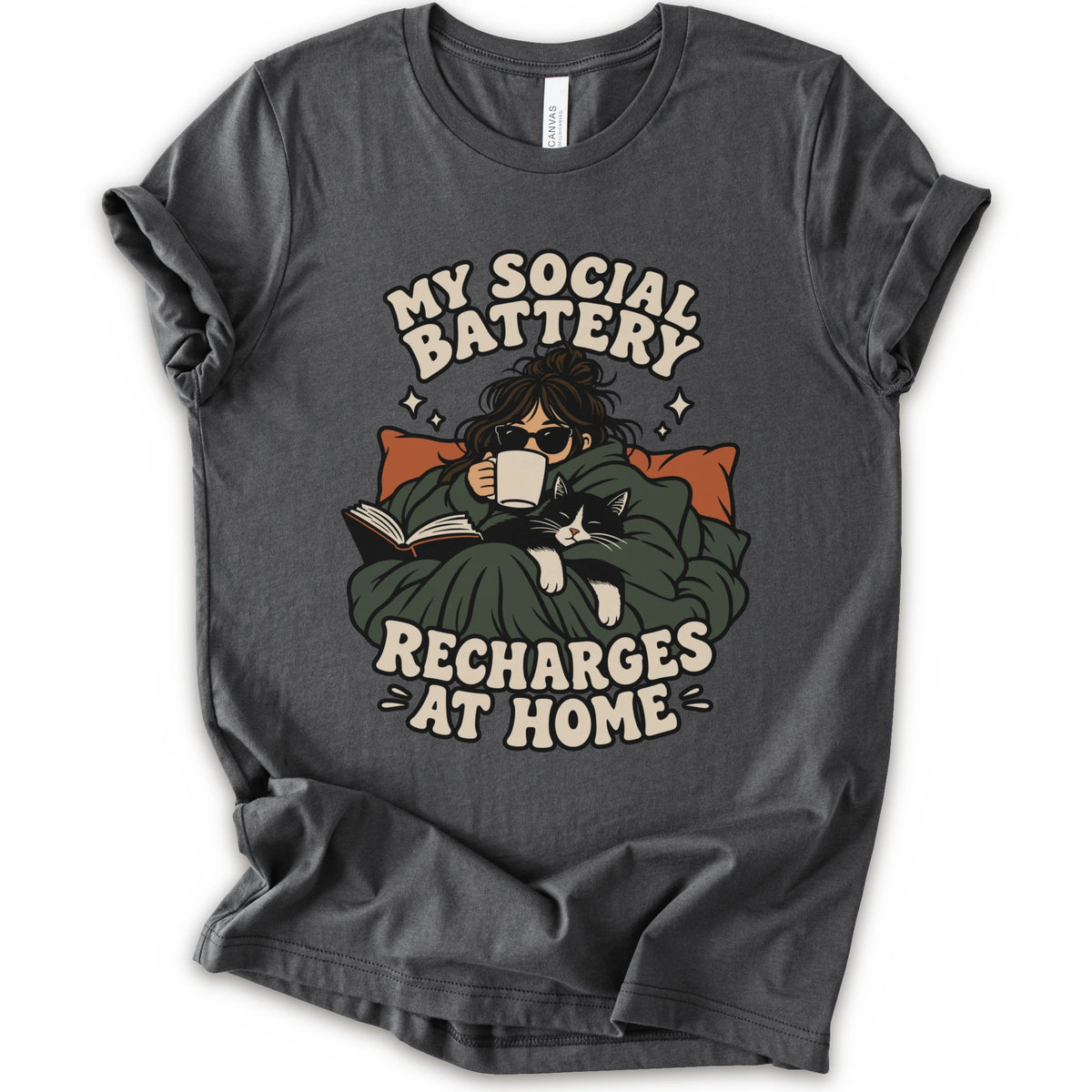 An asphalt short-sleeve graphic t-shirt laid flat against a white background. The graphic features a cozy illustration of a person wrapped in a green blanket reading a book with a black cat, surrounded by the text 'MY SOCIAL BATTERY RECHARGES AT HOME' in a beige retro font.