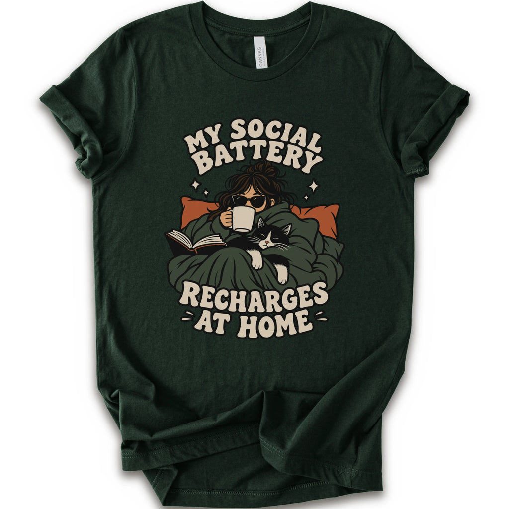 A forest short-sleeve graphic t-shirt features a cozy illustration of a person wrapped in a green blanket reading a book with a black cat, surrounded by the text 'MY SOCIAL BATTERY RECHARGES AT HOME' in a beige retro font.