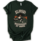 A forest short-sleeve graphic t-shirt features a cozy illustration of a person wrapped in a green blanket reading a book with a black cat, surrounded by the text 'MY SOCIAL BATTERY RECHARGES AT HOME' in a beige retro font.