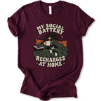A maroon short-sleeve graphic t-shirt features a cozy illustration of a person wrapped in a green blanket reading a book with a black cat, surrounded by the text 'MY SOCIAL BATTERY RECHARGES AT HOME' in a beige retro font.