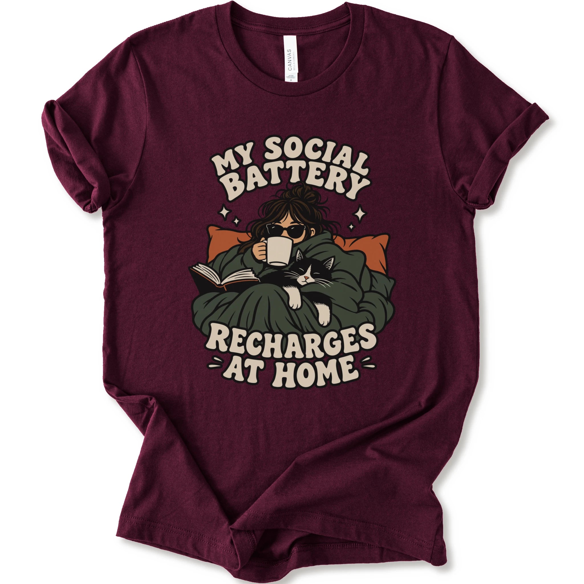 A maroon short-sleeve graphic t-shirt features a cozy illustration of a person wrapped in a green blanket reading a book with a black cat, surrounded by the text 'MY SOCIAL BATTERY RECHARGES AT HOME' in a beige retro font.