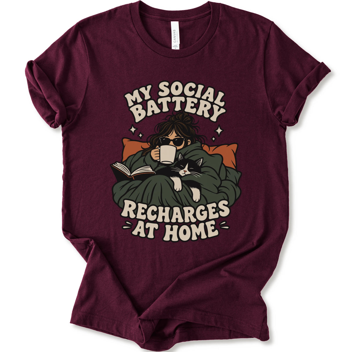 A maroon short-sleeve graphic t-shirt laid flat against a white background. The graphic features a cozy illustration of a person wrapped in a green blanket reading a book with a black cat, surrounded by the text 'MY SOCIAL BATTERY RECHARGES AT HOME' in a beige retro font.