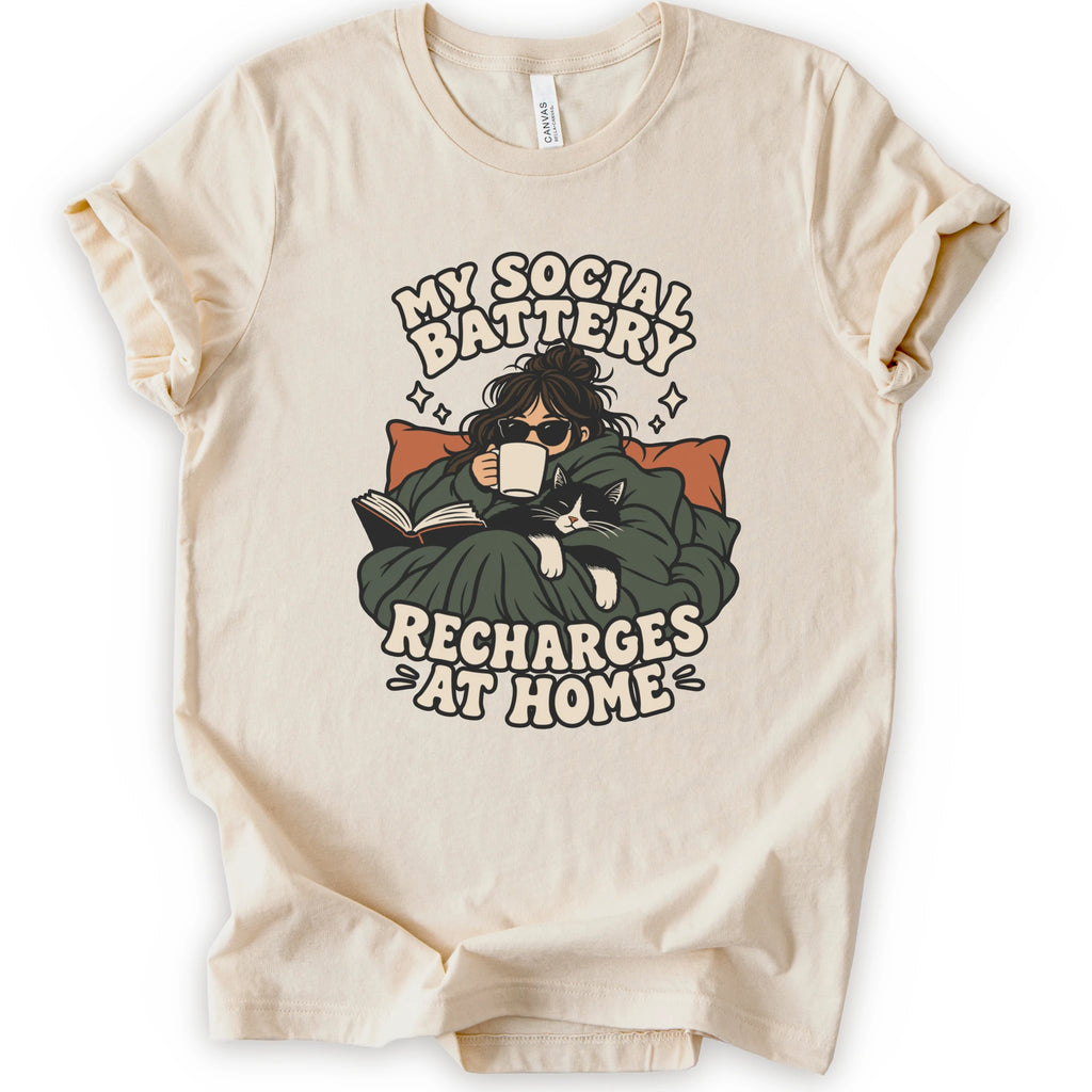 A natural short-sleeve graphic t-shirt features a cozy illustration of a person wrapped in a green blanket reading a book with a black cat, surrounded by the text 'MY SOCIAL BATTERY RECHARGES AT HOME' in a beige retro font.