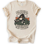 A natural short-sleeve graphic t-shirt features a cozy illustration of a person wrapped in a green blanket reading a book with a black cat, surrounded by the text 'MY SOCIAL BATTERY RECHARGES AT HOME' in a beige retro font.