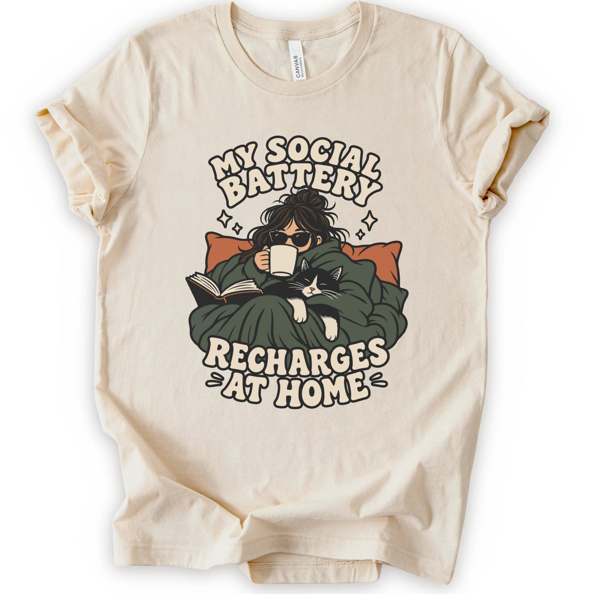A natural short-sleeve graphic t-shirt laid flat against a white background. The graphic features a cozy illustration of a person wrapped in a green blanket reading a book with a black cat, surrounded by the text 'MY SOCIAL BATTERY RECHARGES AT HOME' in a beige retro font.