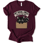 A maroon short-sleeve crewneck t-shirt with a graphic print of a black cat in a cardboard box and the text 'MY SOCIAL CIRCLE FITS IN THIS BOX."