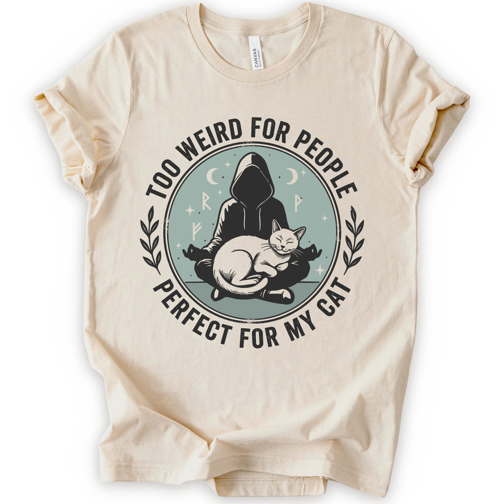 A natural short-sleeve crewneck t-shirt with a circular graphic on the front. The graphic features a dark silhouette of a person in a hood sitting cross-legged next to a white cat, surrounded by the text 'TOO WEIRD FOR PEOPLE' on top and 'PERFECT FOR MY CAT' on the bottom, with small moon and star details.