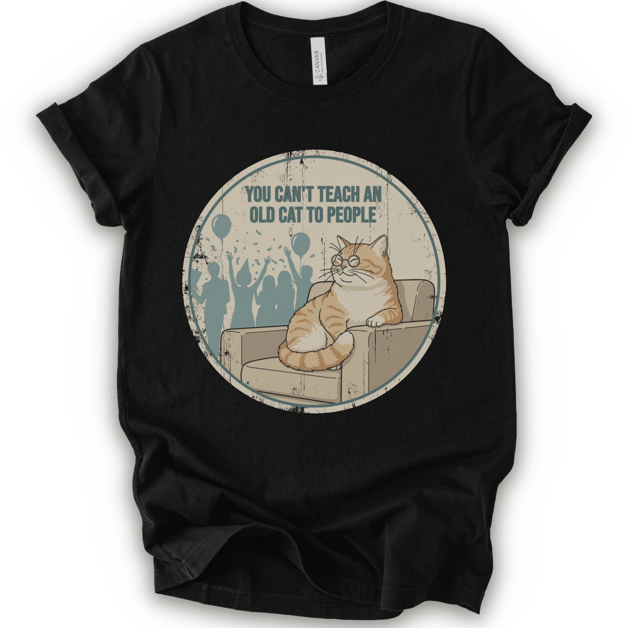 A black short-sleeve crewneck t-shirt with a circular graphic print on the front. The graphic features an orange tabby cat sitting on a beige armchair inside a distressed circle, with the text 'YOU CAN'T TEACH AN OLD CAT TO PEOPLE' above it and blue silhouettes of people in the background.