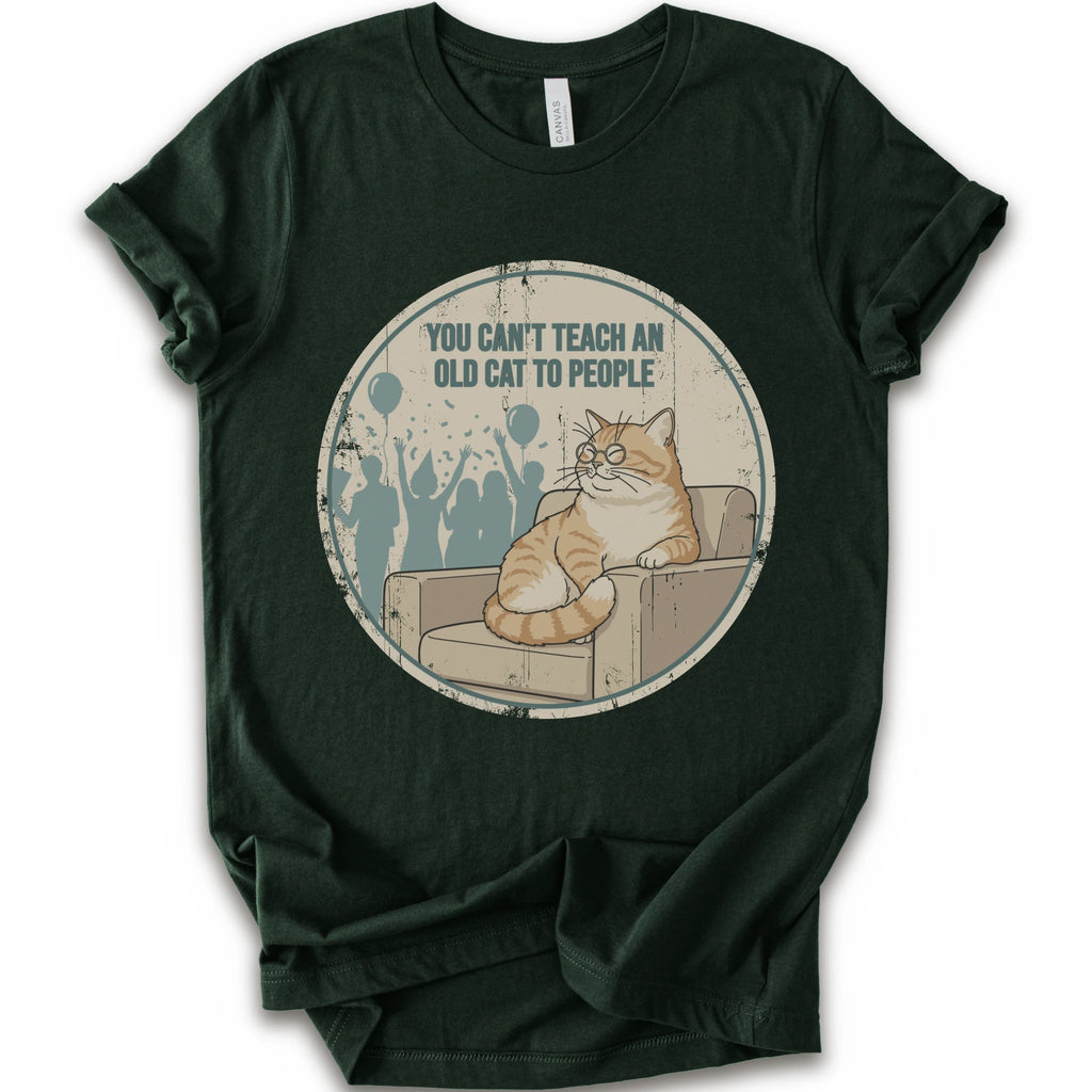 A forest short-sleeve crewneck t-shirt with a circular graphic print on the front. The graphic features an orange tabby cat sitting on a beige armchair inside a distressed circle, with the text 'YOU CAN'T TEACH AN OLD CAT TO PEOPLE' above it and blue silhouettes of people in the background.