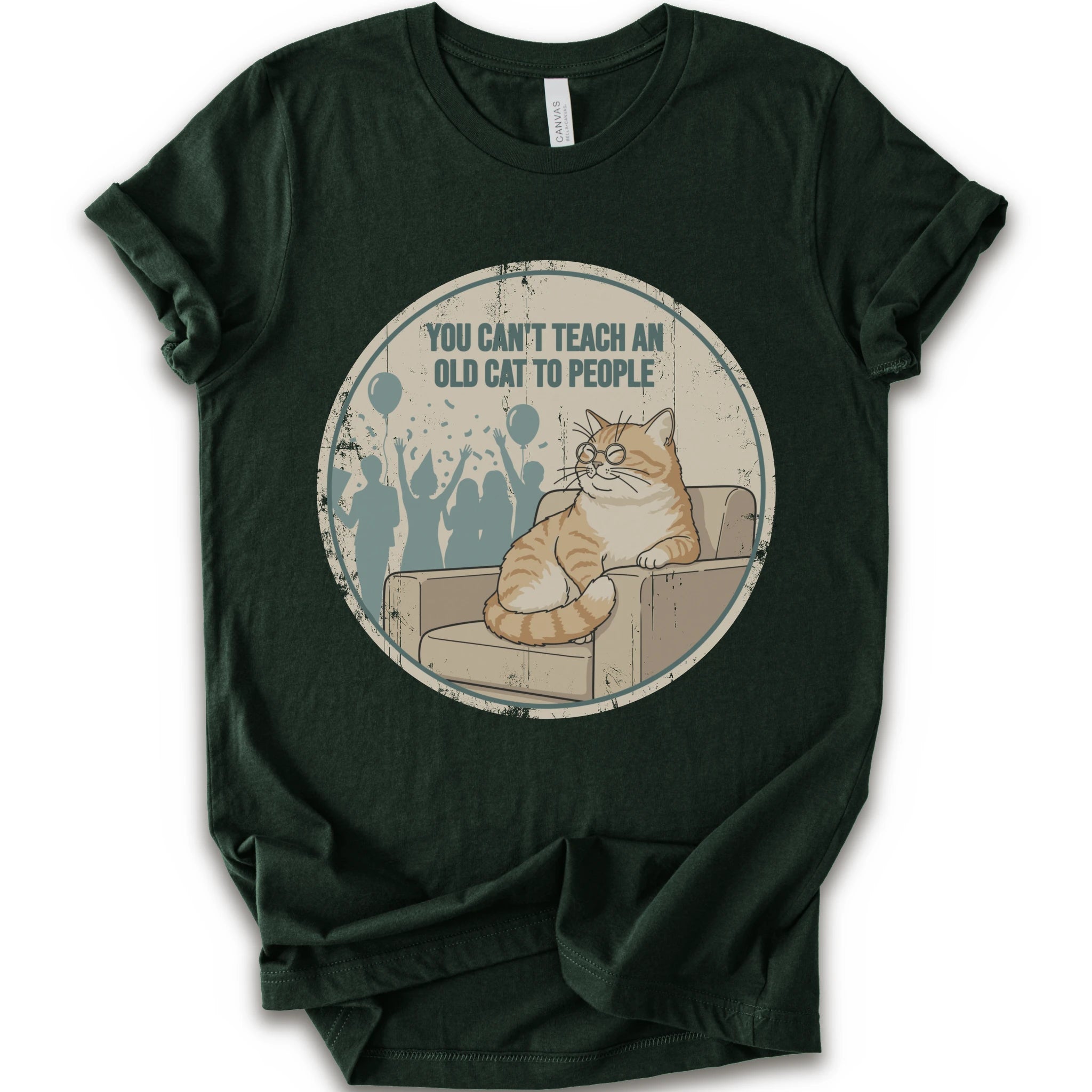 A forest short-sleeve crewneck t-shirt with a circular graphic print on the front. The graphic features an orange tabby cat sitting on a beige armchair inside a distressed circle, with the text 'YOU CAN'T TEACH AN OLD CAT TO PEOPLE' above it and blue silhouettes of people in the background.