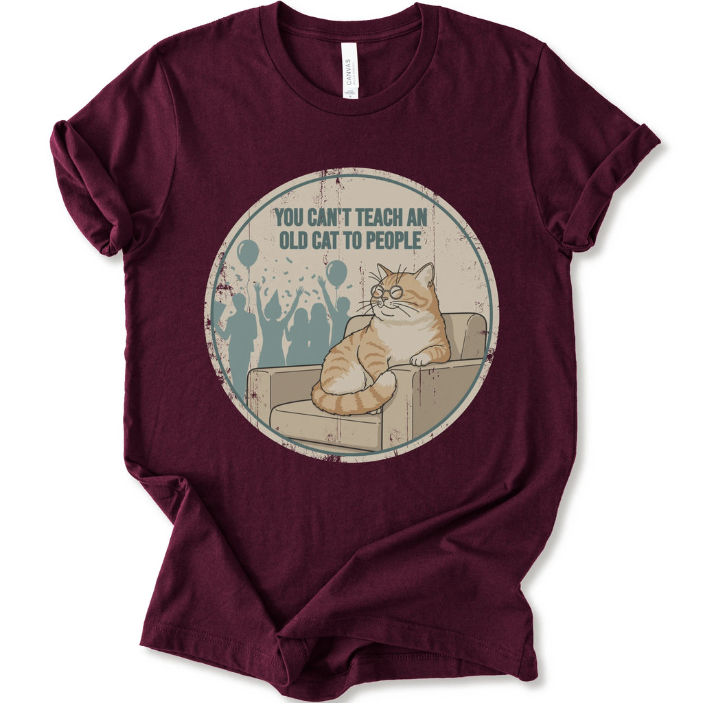 A maroon short-sleeve crewneck t-shirt with a circular graphic print on the front. The graphic features an orange tabby cat sitting on a beige armchair inside a distressed circle, with the text 'YOU CAN'T TEACH AN OLD CAT TO PEOPLE' above it and blue silhouettes of people in the background.