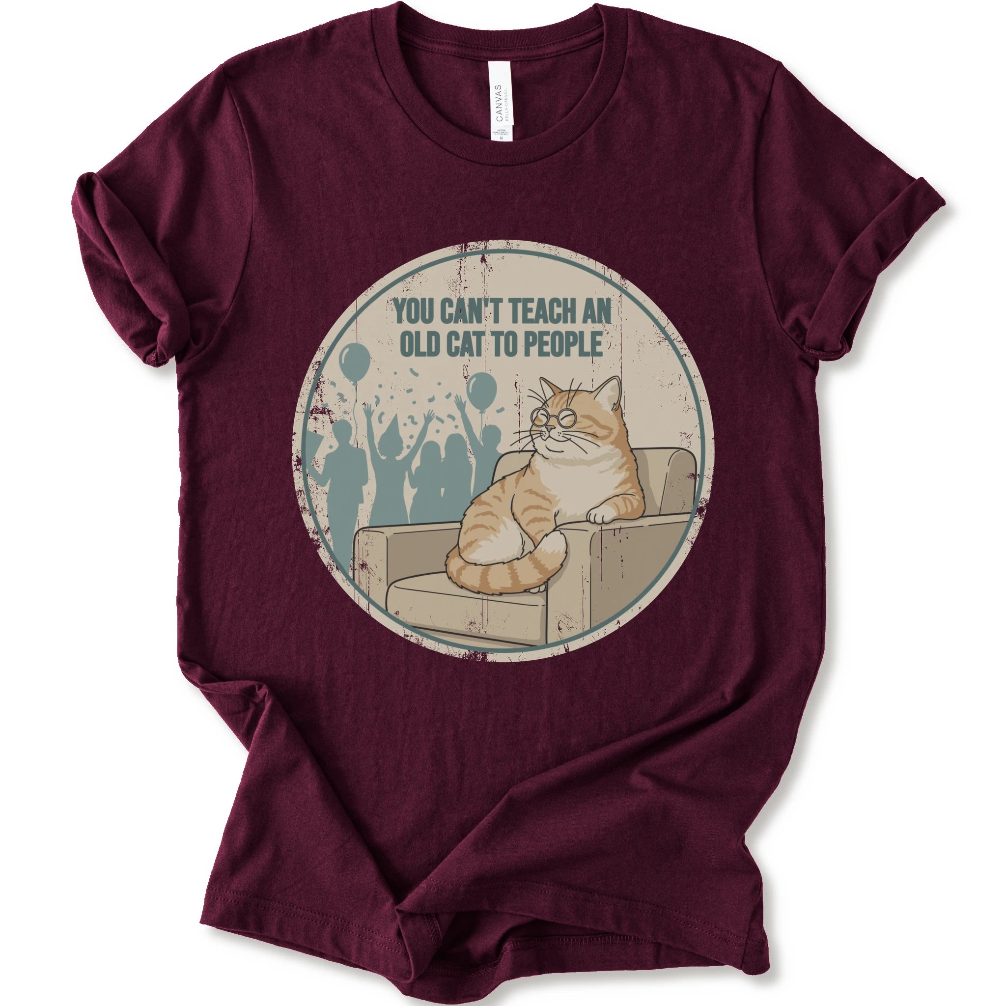 A maroon short-sleeve crewneck t-shirt with a circular graphic print on the front. The graphic features an orange tabby cat sitting on a beige armchair inside a distressed circle, with the text 'YOU CAN'T TEACH AN OLD CAT TO PEOPLE' above it and blue silhouettes of people in the background.