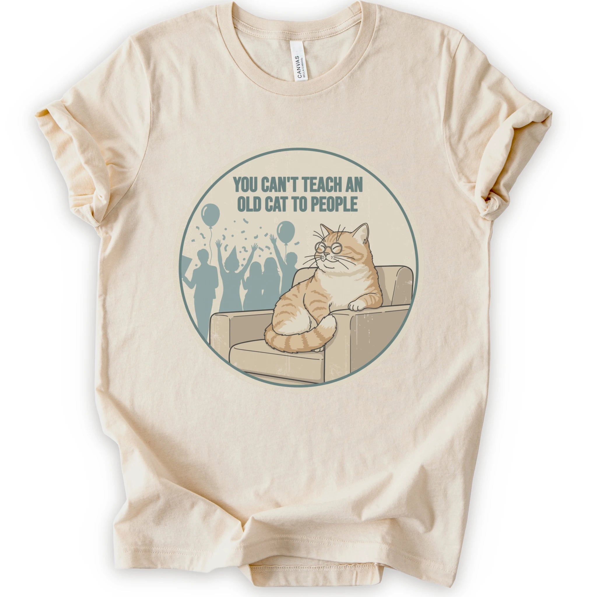 A natural short-sleeve crewneck t-shirt with a circular graphic print on the front. The graphic features an orange tabby cat sitting on a beige armchair inside a distressed circle, with the text 'YOU CAN'T TEACH AN OLD CAT TO PEOPLE' above it and blue silhouettes of people in the background.
