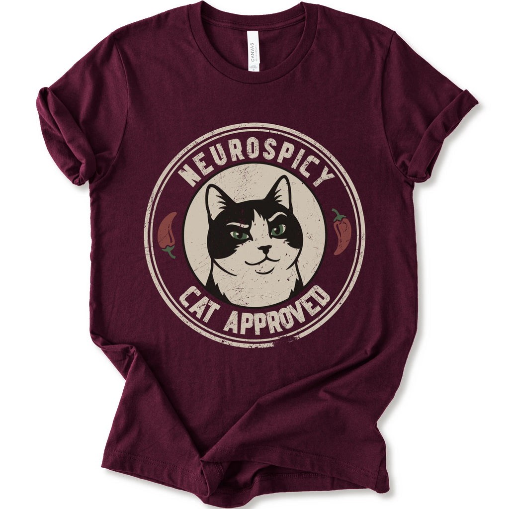 A maroon short-sleeve crewneck t-shirt with a distressed circular graphic on the chest featuring a black and white cat with green eyes, red peppers, and the text 'NEUROSPICY, CAT APPROVED'.