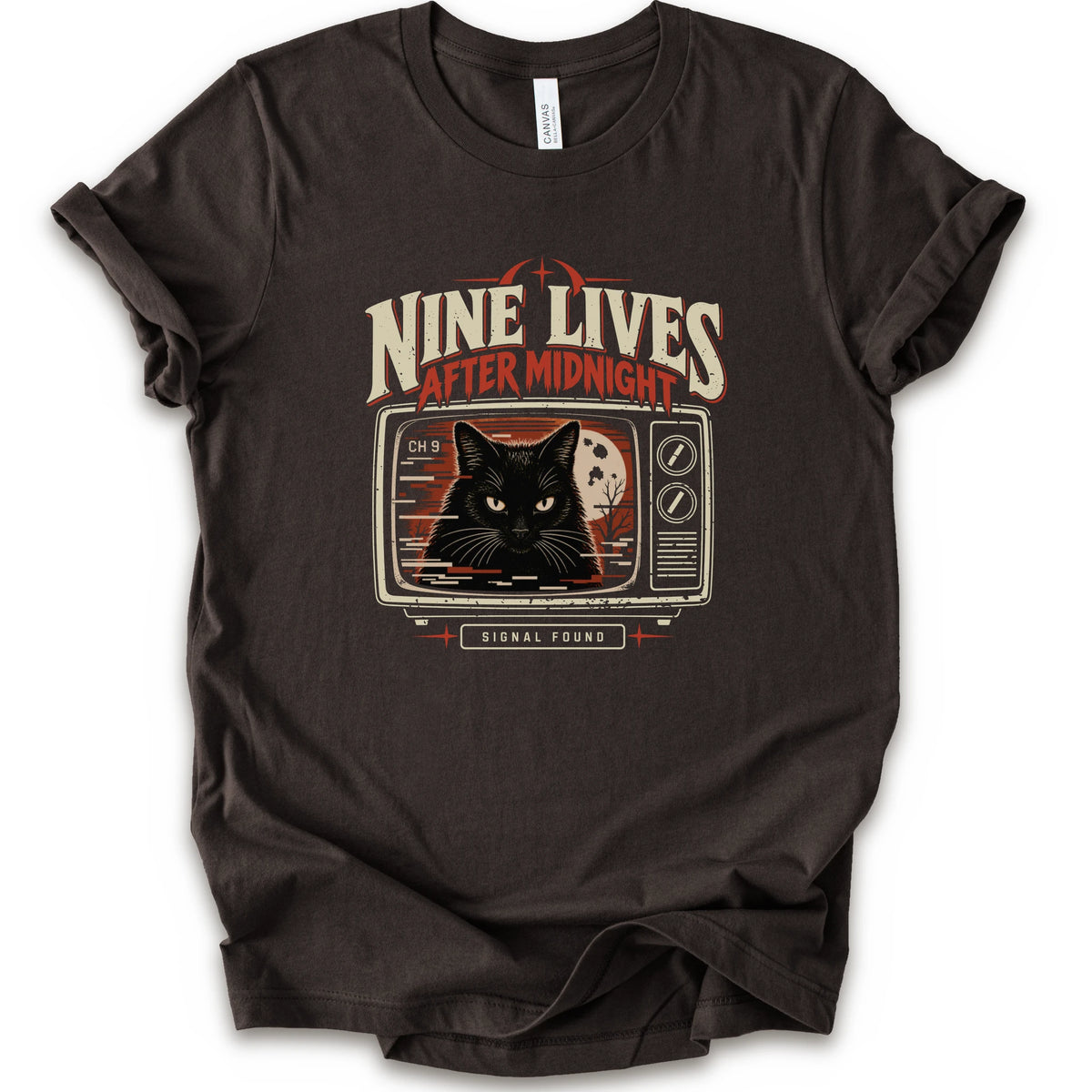 A brown short-sleeve crewneck t-shirt with a vintage-style graphic on the front. The graphic features the text 'NINE LIVES AFTER MIDNIGHT' in distressed cream and red lettering above an illustration of a black cat's face on a retro television screen, with 'SIGNAL FOUND' written below.