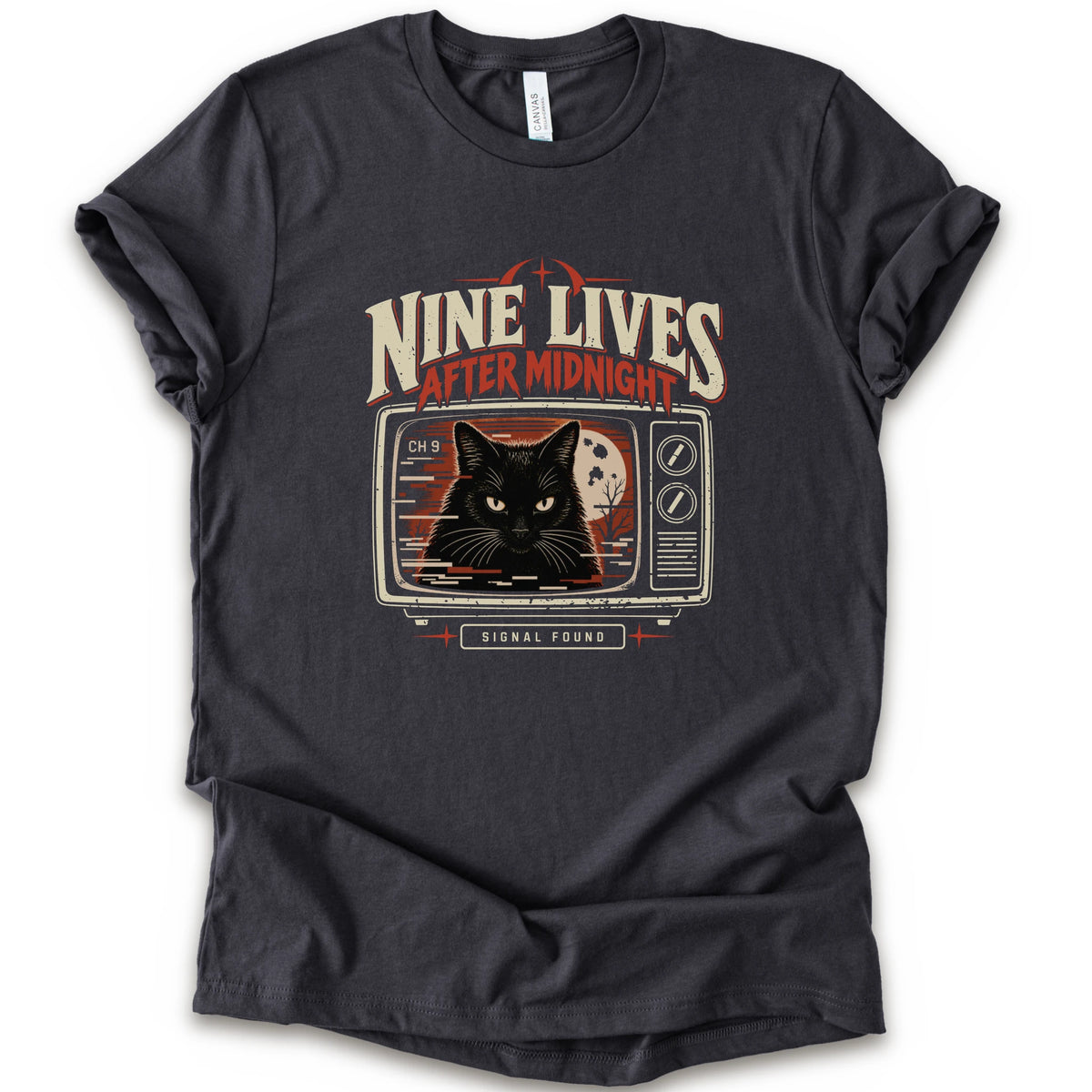 A dark grey short-sleeve crewneck t-shirt with a vintage-style graphic on the front. The graphic features the text 'NINE LIVES AFTER MIDNIGHT' in distressed cream and red lettering above an illustration of a black cat's face on a retro television screen, with 'SIGNAL FOUND' written below.
