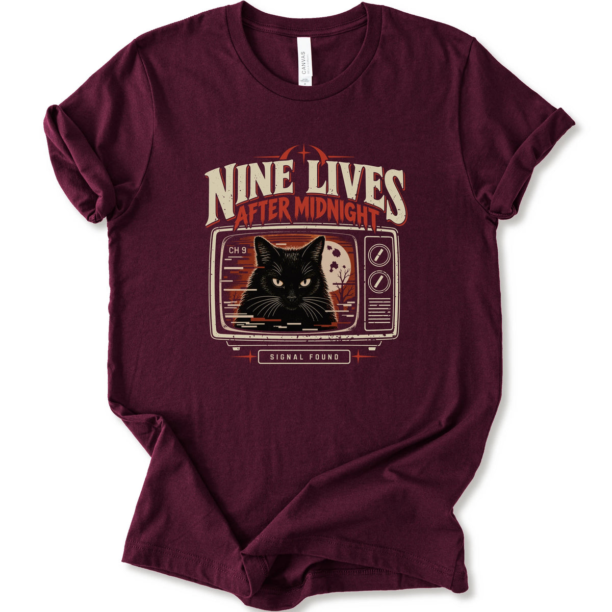 A maroon short-sleeve crewneck t-shirt with a vintage-style graphic on the front. The graphic features the text 'NINE LIVES AFTER MIDNIGHT' in distressed cream and red lettering above an illustration of a black cat's face on a retro television screen, with 'SIGNAL FOUND' written below.