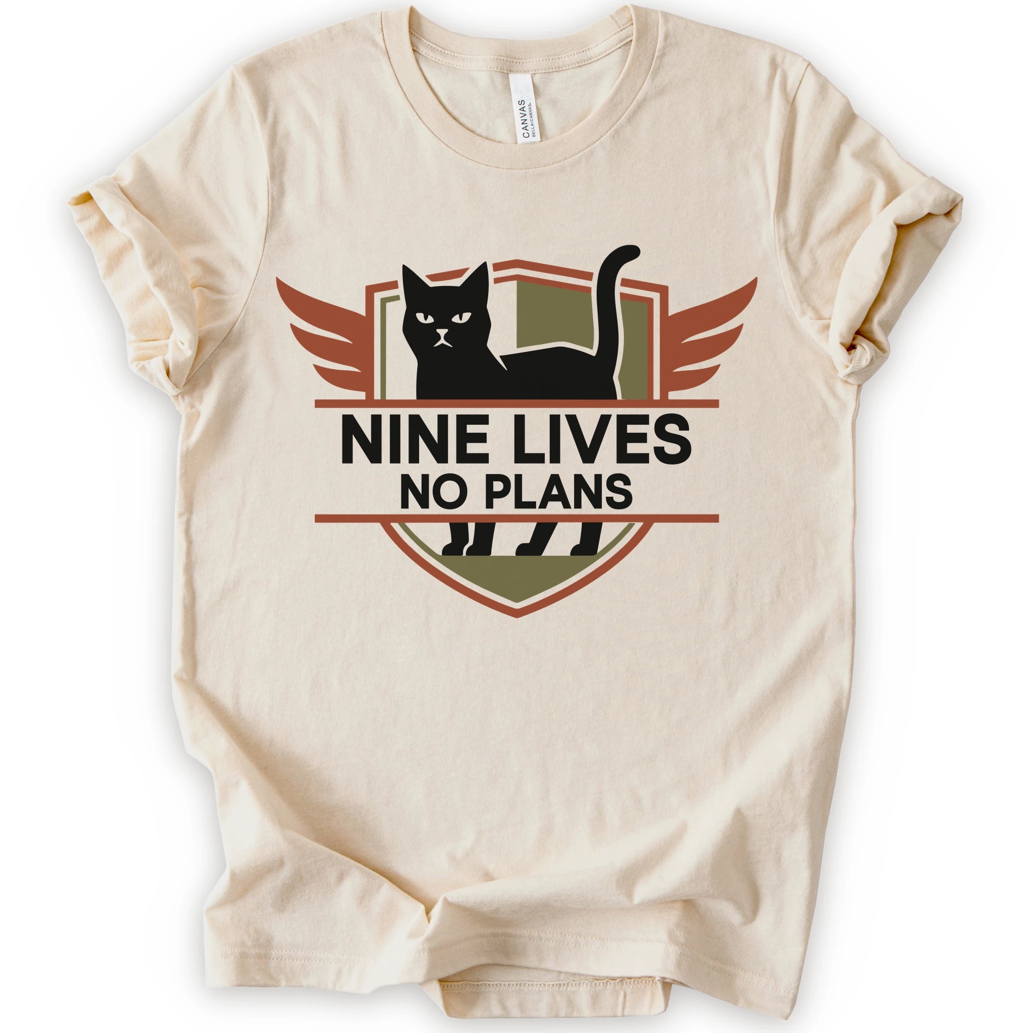A natural T-shirt with a design that reads “NINE LIVES NO PLANS” beneath a black cat centered inside a winged retro badge. Muted rust orange and olive shapes create a dark vintage-style emblem.
