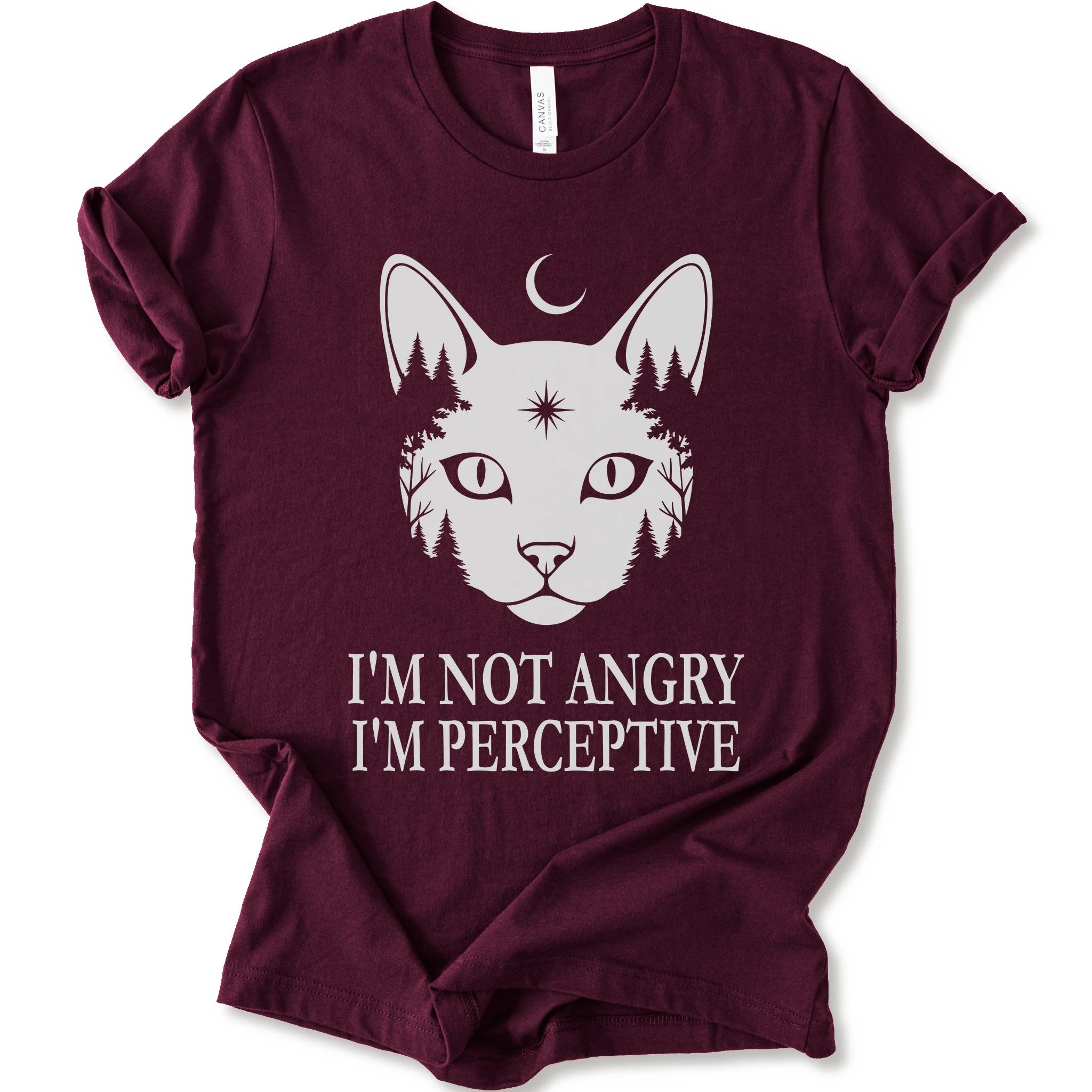 A maroon short-sleeve crewneck t-shirt with a white graphic of a cat's face containing pine trees and a crescent moon, with the text 'I'M NOT ANGRY I'M PERCEPTIVE' printed below.