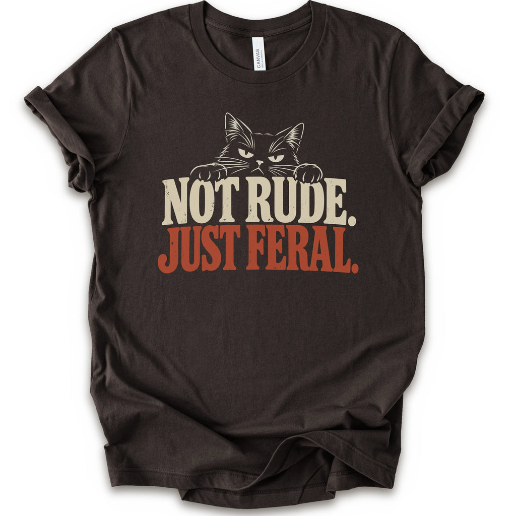 A brown short-sleeve t-shirt with a graphic print of a cat with narrowed eyes and visible paws peers over the words “NOT RUDE.” above a large rust-colored “JUST FERAL.” The design uses bold distressed serif lettering in cream and rust with a retro graphic look.