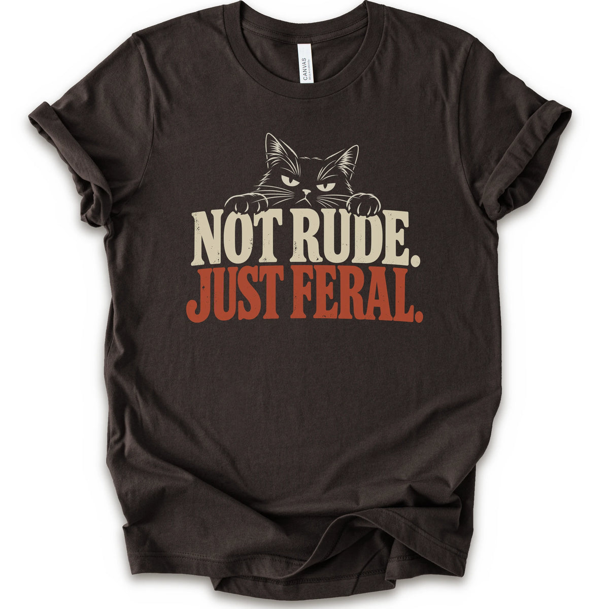 A brown short-sleeve t-shirt with a graphic print of a cat with narrowed eyes and visible paws peers over the words “NOT RUDE.” above a large rust-colored “JUST FERAL.” The design uses bold distressed serif lettering in cream and rust with a retro graphic look.