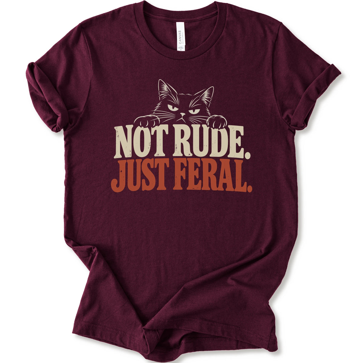 A maroon short-sleeve t-shirt with a graphic print of a cat with narrowed eyes and visible paws peers over the words “NOT RUDE.” above a large rust-colored “JUST FERAL.” The design uses bold distressed serif lettering in cream and rust with a retro graphic look.
