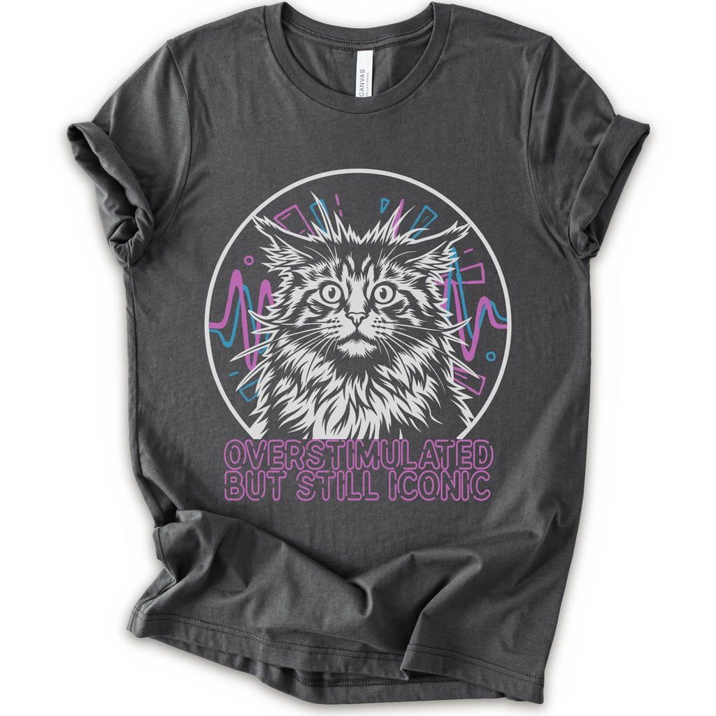 An asphalt short-sleeve crewneck t-shirt featuring a graphic of a white and gray maine con cat inside a circular frame with pink and blue accents, and the text 'OVERSTIMULATED BUT STILL ICONIC' in pink below.