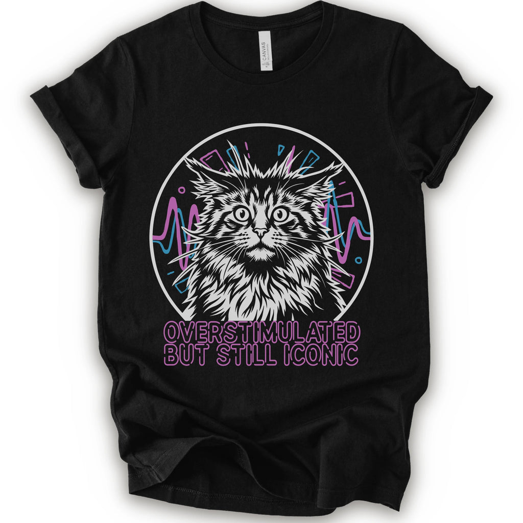 A black short-sleeve crewneck t-shirt featuring a graphic of a white and gray maine con cat inside a circular frame with pink and blue accents, and the text 'OVERSTIMULATED BUT STILL ICONIC' in pink below.