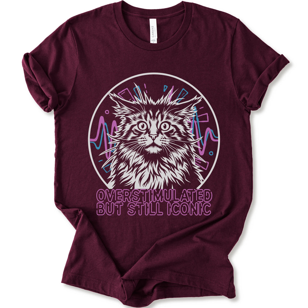 A maroon short-sleeve crewneck t-shirt featuring a graphic of a white and gray maine con cat inside a circular frame with pink and blue accents, and the text 'OVERSTIMULATED BUT STILL ICONIC' in pink below.