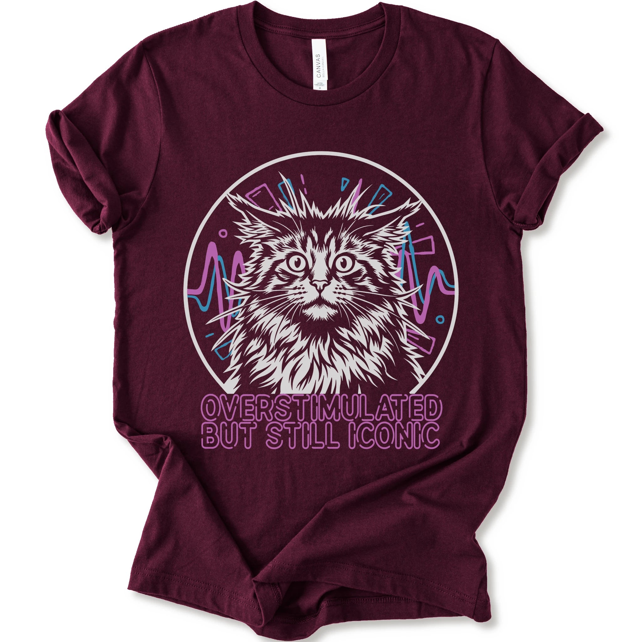 A maroon short-sleeve crewneck t-shirt featuring a graphic of a white and gray maine con cat inside a circular frame with pink and blue accents, and the text 'OVERSTIMULATED BUT STILL ICONIC' in pink below.