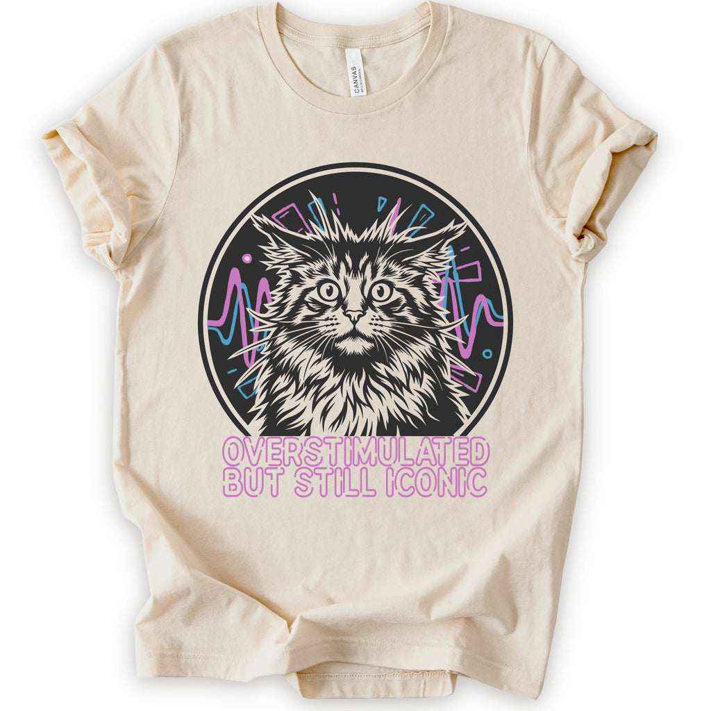 A natural short-sleeve crewneck t-shirt featuring a graphic of a white and gray maine con cat inside a circular frame with pink and blue accents, and the text 'OVERSTIMULATED BUT STILL ICONIC' in pink below.