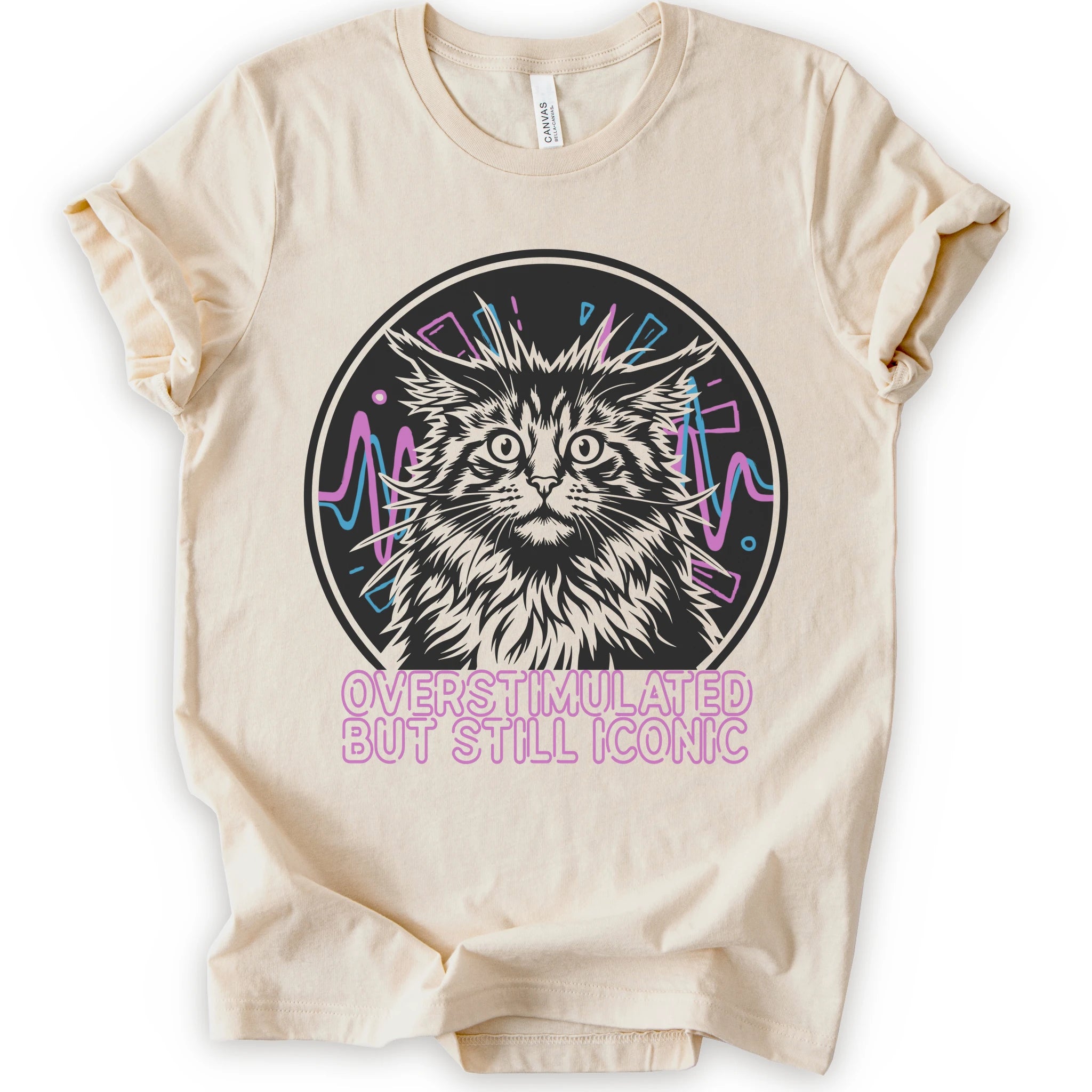 A natural short-sleeve crewneck t-shirt featuring a graphic of a white and gray maine con cat inside a circular frame with pink and blue accents, and the text 'OVERSTIMULATED BUT STILL ICONIC' in pink below.