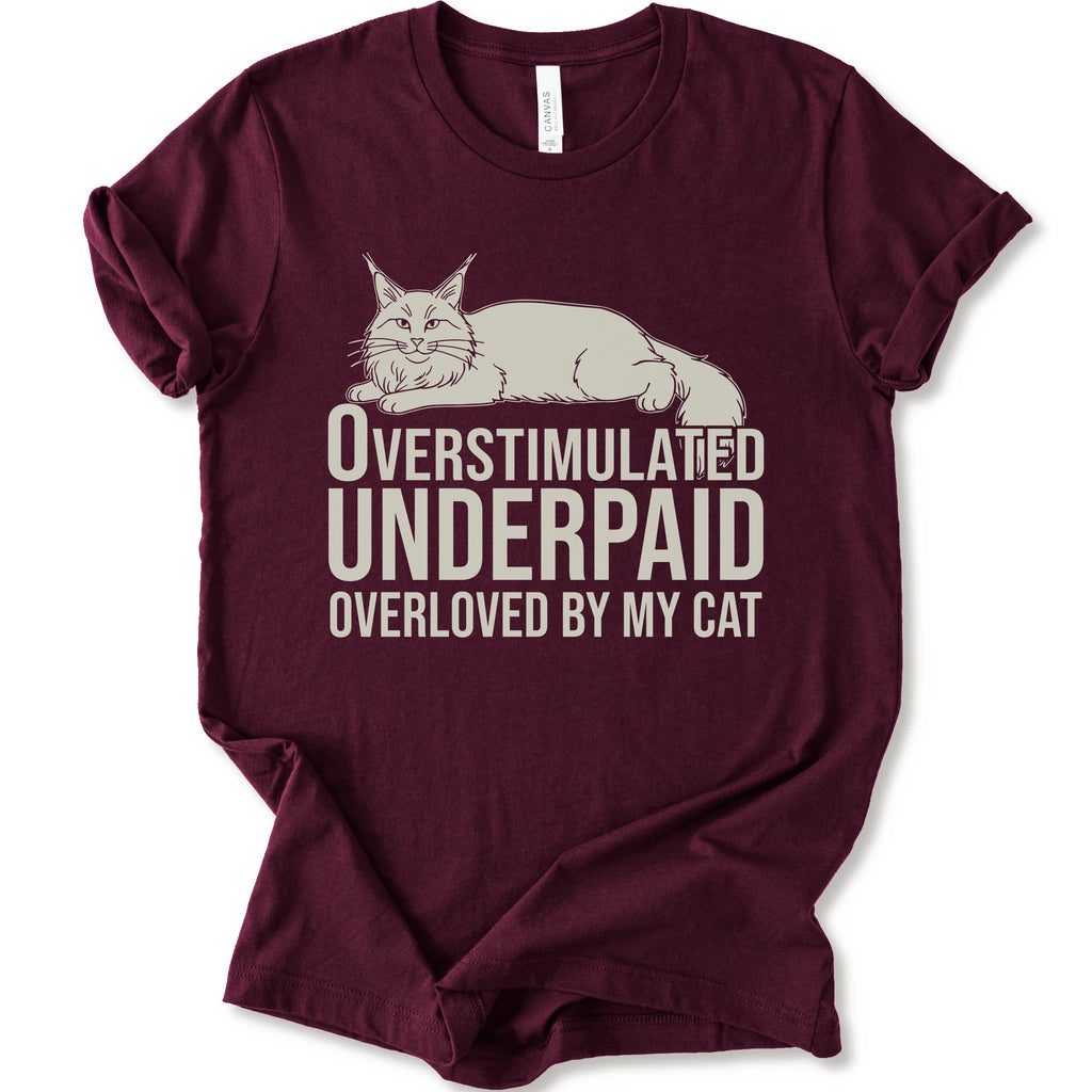 A maroon short-sleeve crewneck T-shirt with a lounging cat graphic and “Overstimulated, Underpaid, Overloved by My Cat” text.
