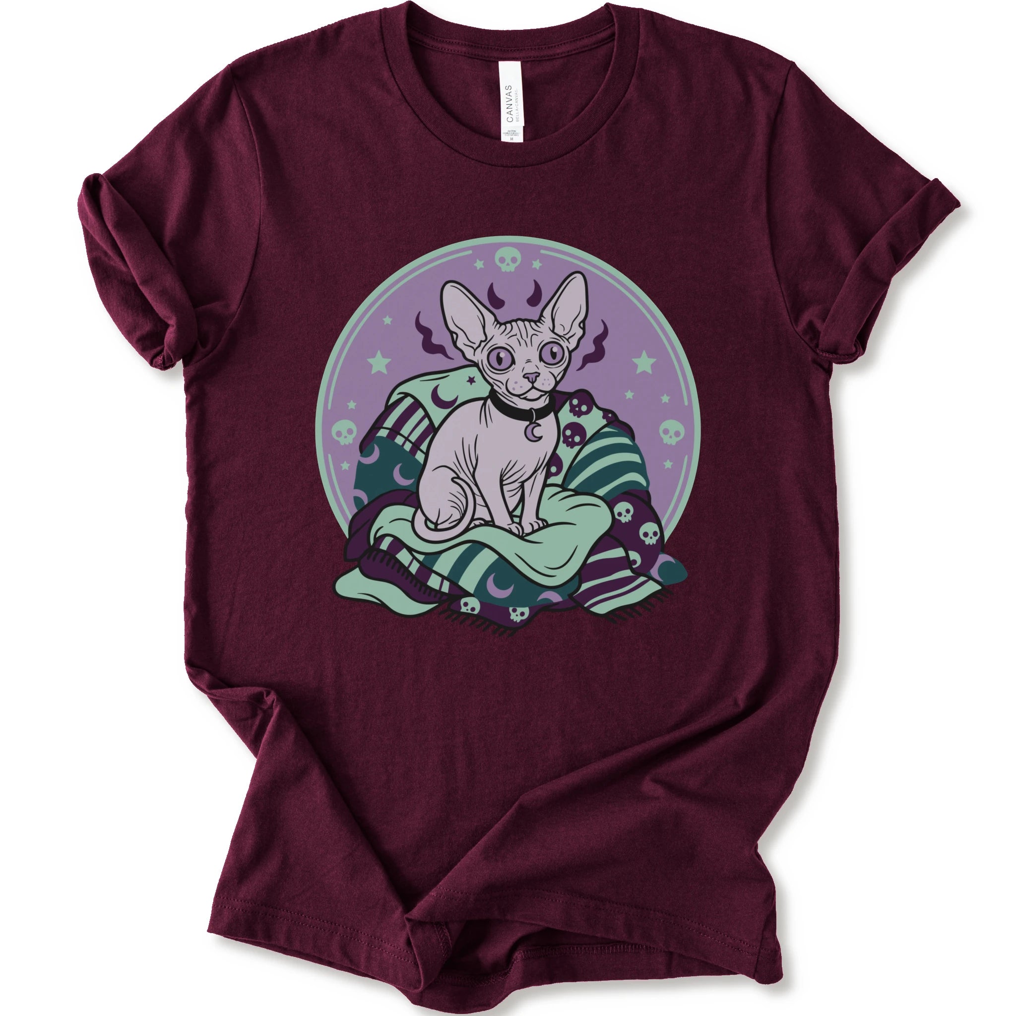 A maroon short-sleeve crewneck t-shirt featuring a graphic print of a hairless cat sitting on a green and purple patterned blanket inside a purple circular frame.