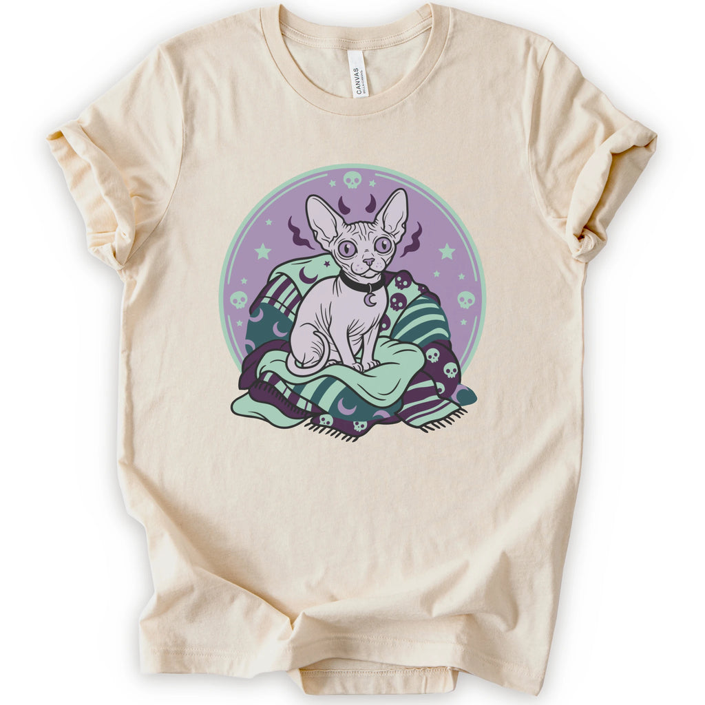 A natural short-sleeve crewneck t-shirt featuring a graphic print of a hairless cat sitting on a green and purple patterned blanket inside a purple circular frame.
