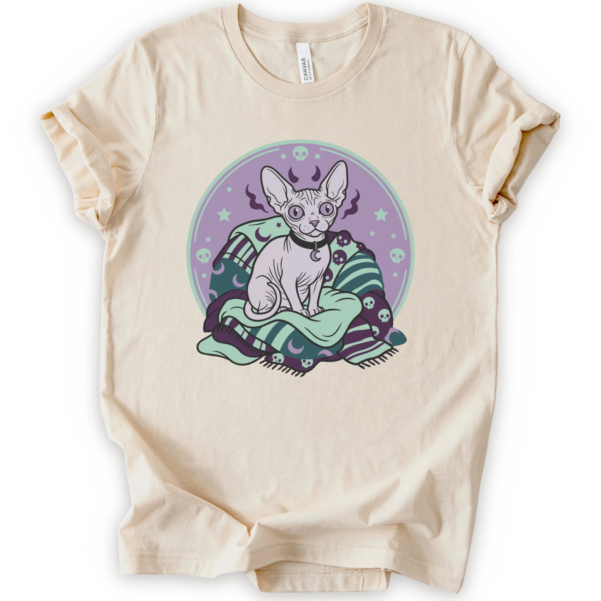 A natural short-sleeve crewneck t-shirt featuring a graphic print of a hairless cat sitting on a green and purple patterned blanket inside a purple circular frame.