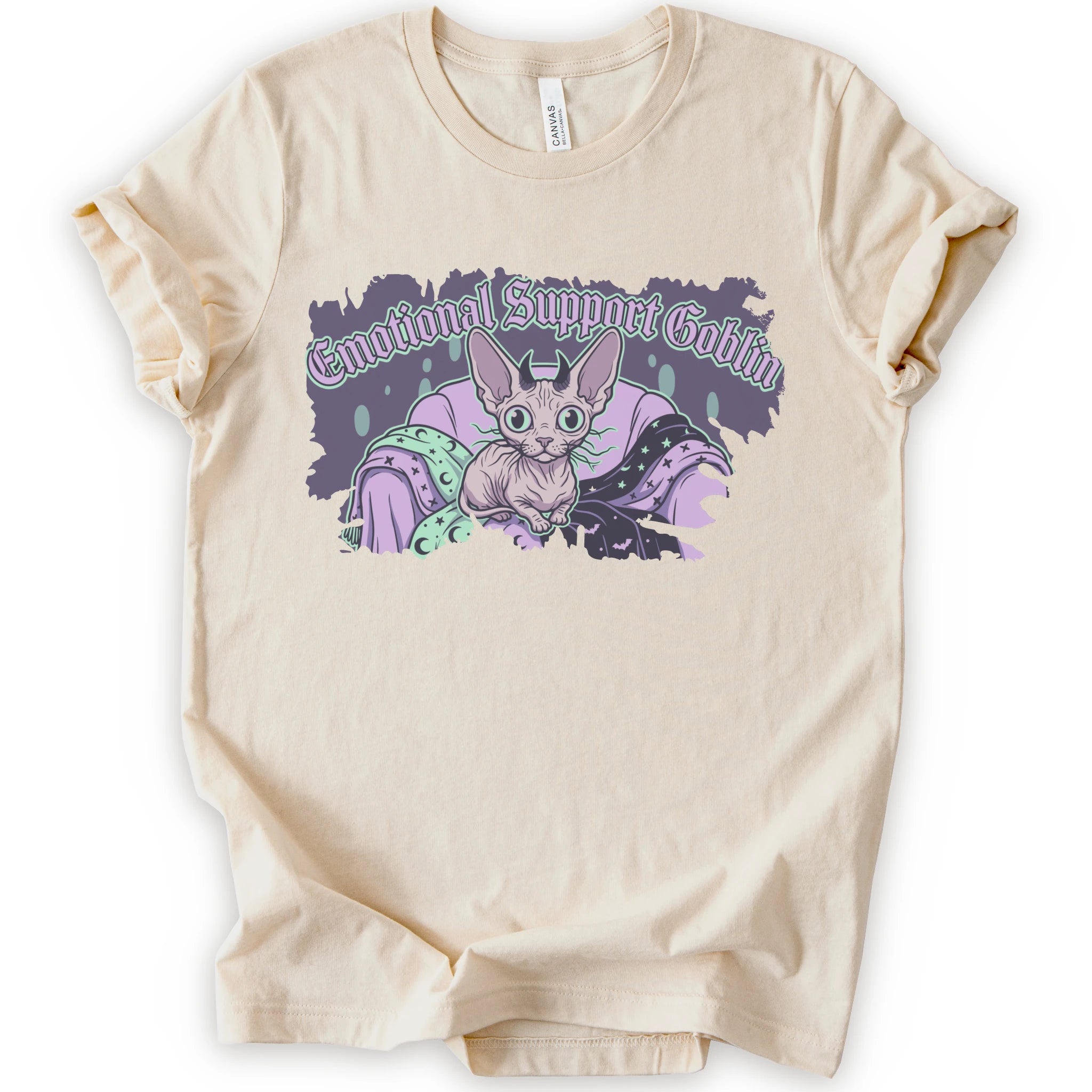 A natural short-sleeve t-shirt with a graphic print of a cartoon sphynx cat in a purple blanket with the text 'Emotional Support Goblin'.
