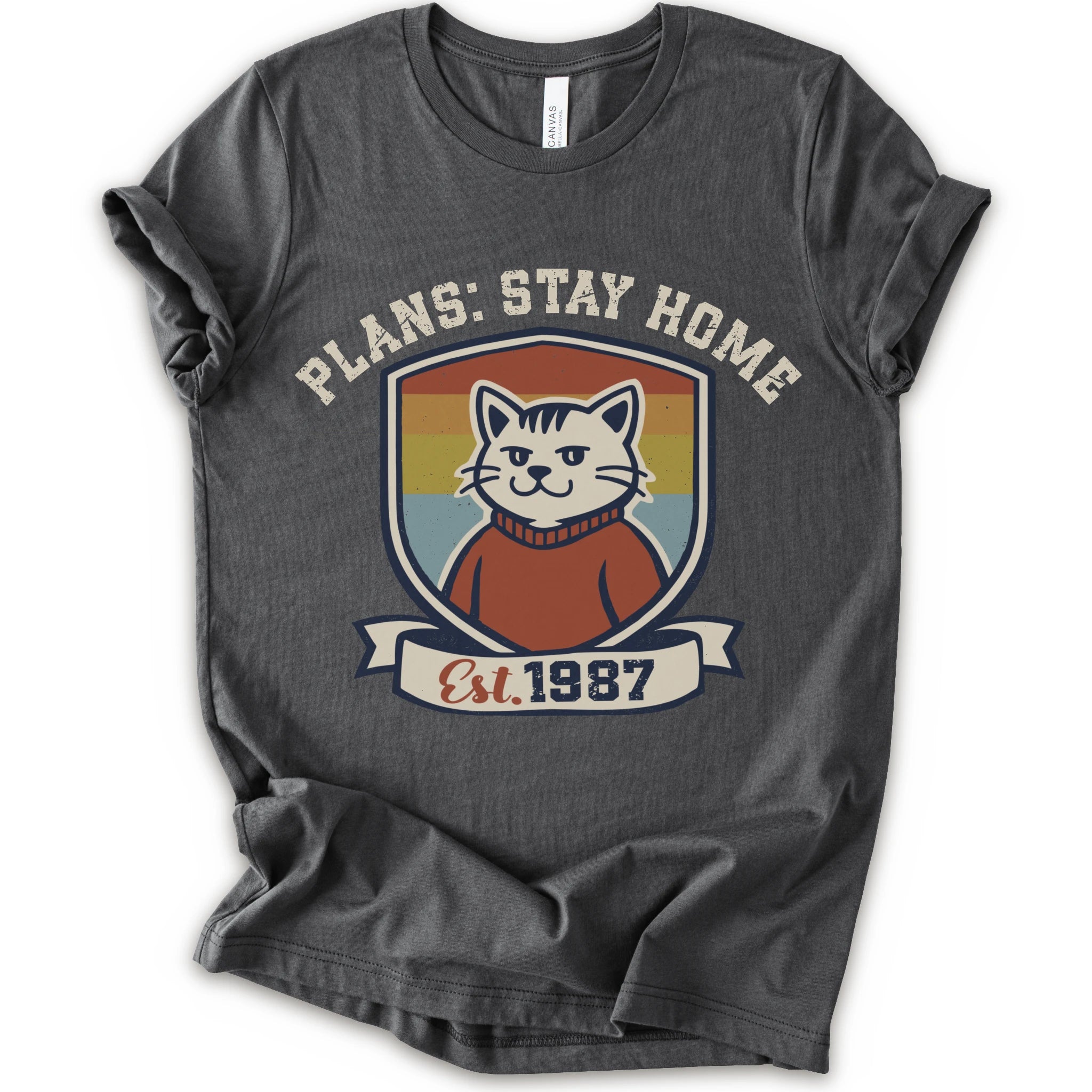 A asphalt short-sleeve crewneck t-shirt with a retro-style graphic of a smiling cat in a red sweater inside a shield, with the text 'PLANS: STAY HOME' and 'Est. 1987' on a banner below.