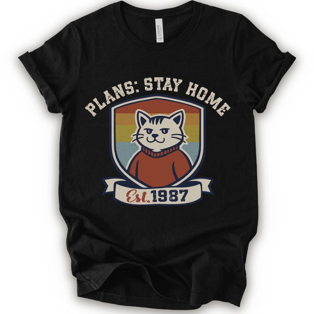 A black short-sleeve crewneck t-shirt with a retro-style graphic of a smiling cat in a red sweater inside a shield, with the text 'PLANS: STAY HOME' and 'Est. 1987' on a banner below.