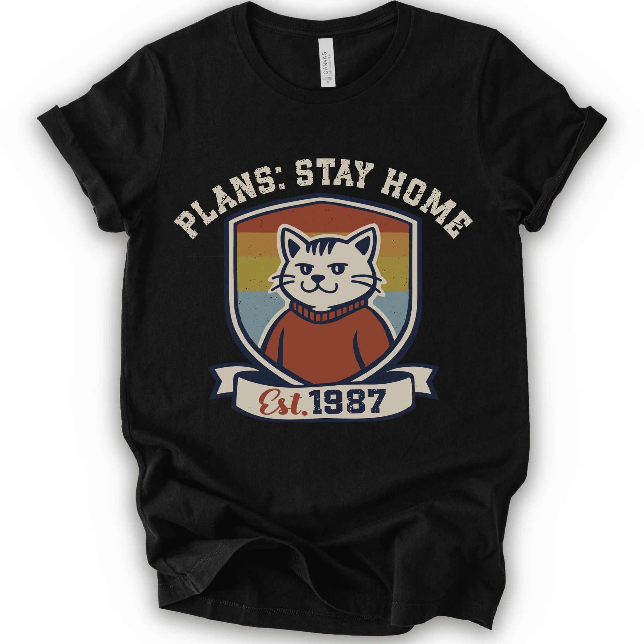 A black short-sleeve crewneck t-shirt with a retro-style graphic of a smiling cat in a red sweater inside a shield, with the text 'PLANS: STAY HOME' and 'Est. 1987' on a banner below.
