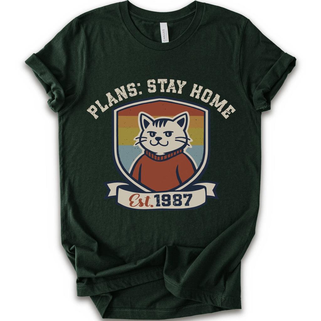 A forest short-sleeve crewneck t-shirt with a retro-style graphic of a smiling cat in a red sweater inside a shield, with the text 'PLANS: STAY HOME' and 'Est. 1987' on a banner below.
