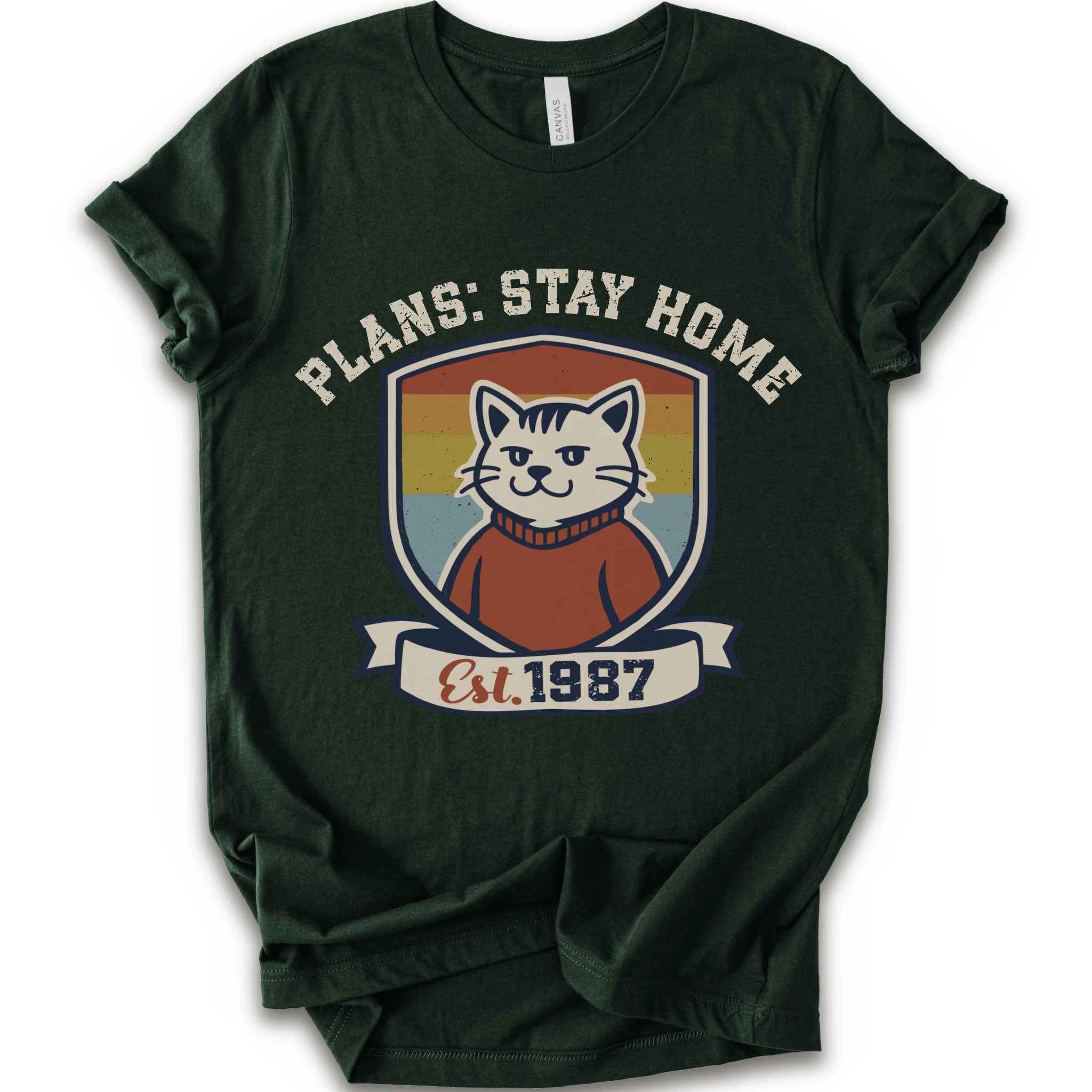 A forest short-sleeve crewneck t-shirt with a retro-style graphic of a smiling cat in a red sweater inside a shield, with the text 'PLANS: STAY HOME' and 'Est. 1987' on a banner below.