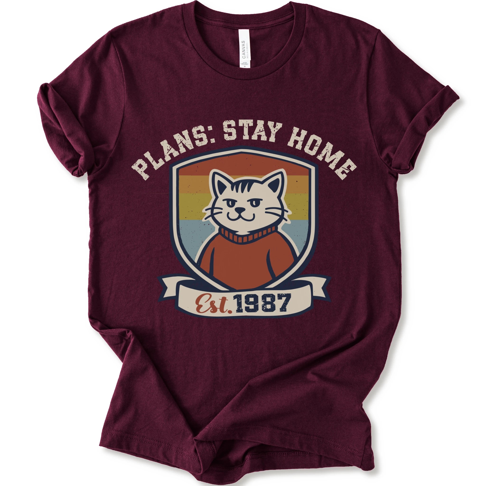 A maroon short-sleeve crewneck t-shirt with a retro-style graphic of a smiling cat in a red sweater inside a shield, with the text 'PLANS: STAY HOME' and 'Est. 1987' on a banner below.