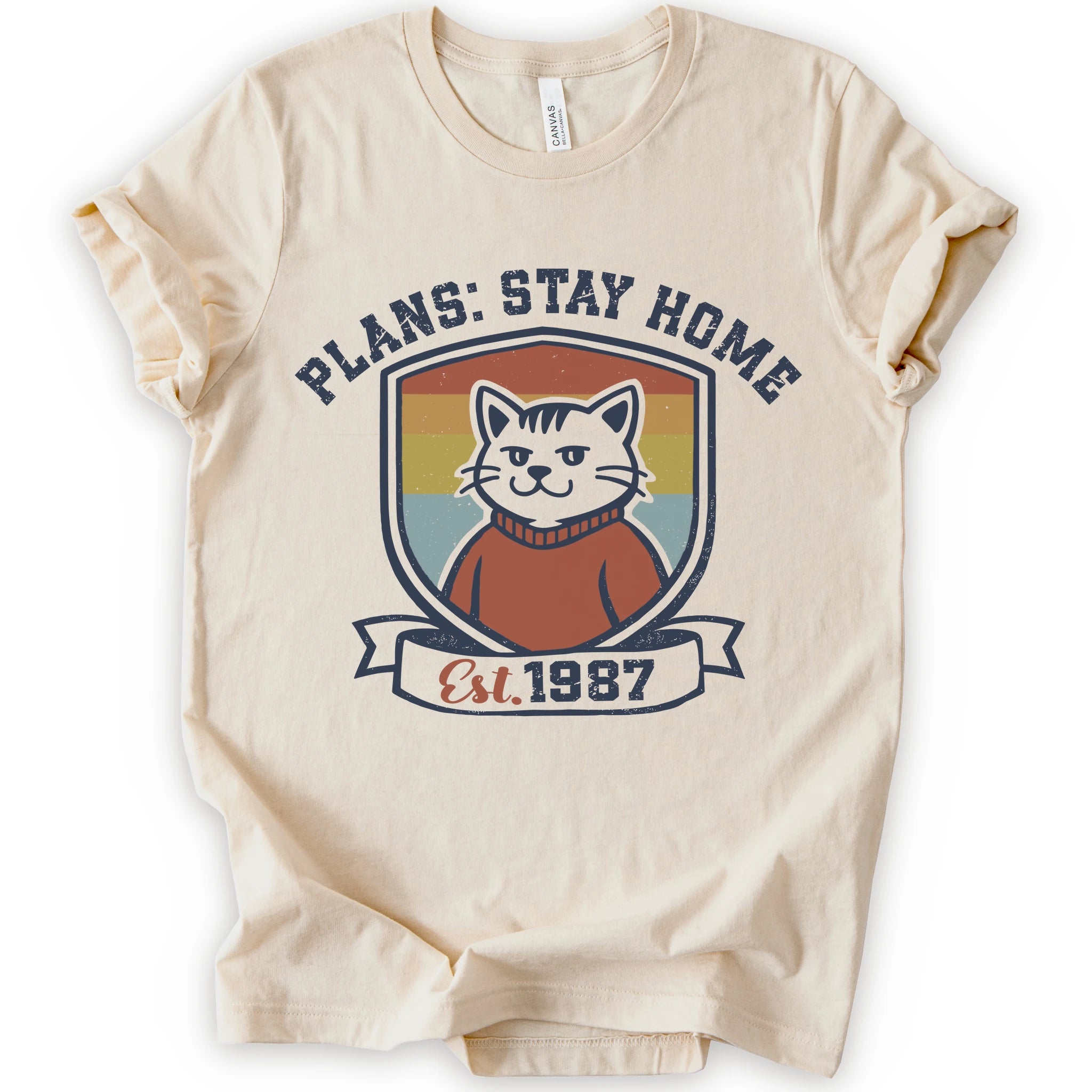 A natural short-sleeve crewneck t-shirt with a retro-style graphic of a smiling cat in a red sweater inside a shield, with the text 'PLANS: STAY HOME' and 'Est. 1987' on a banner below.