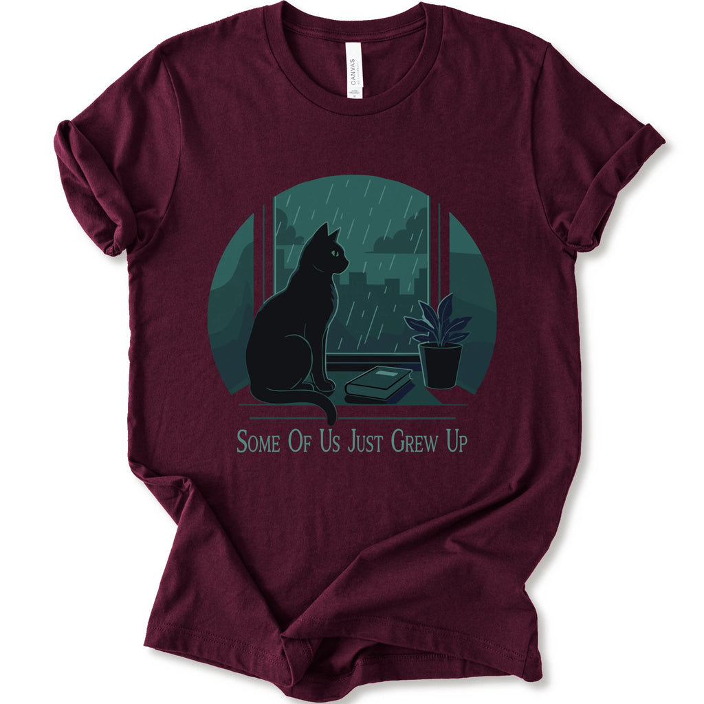 A maroon short-sleeve crewneck t-shirt with a graphic print of a black cat sitting by a window in the rain, next to a small potted plant, with the text 'SOME OF US JUST GREW UP' printed below.
