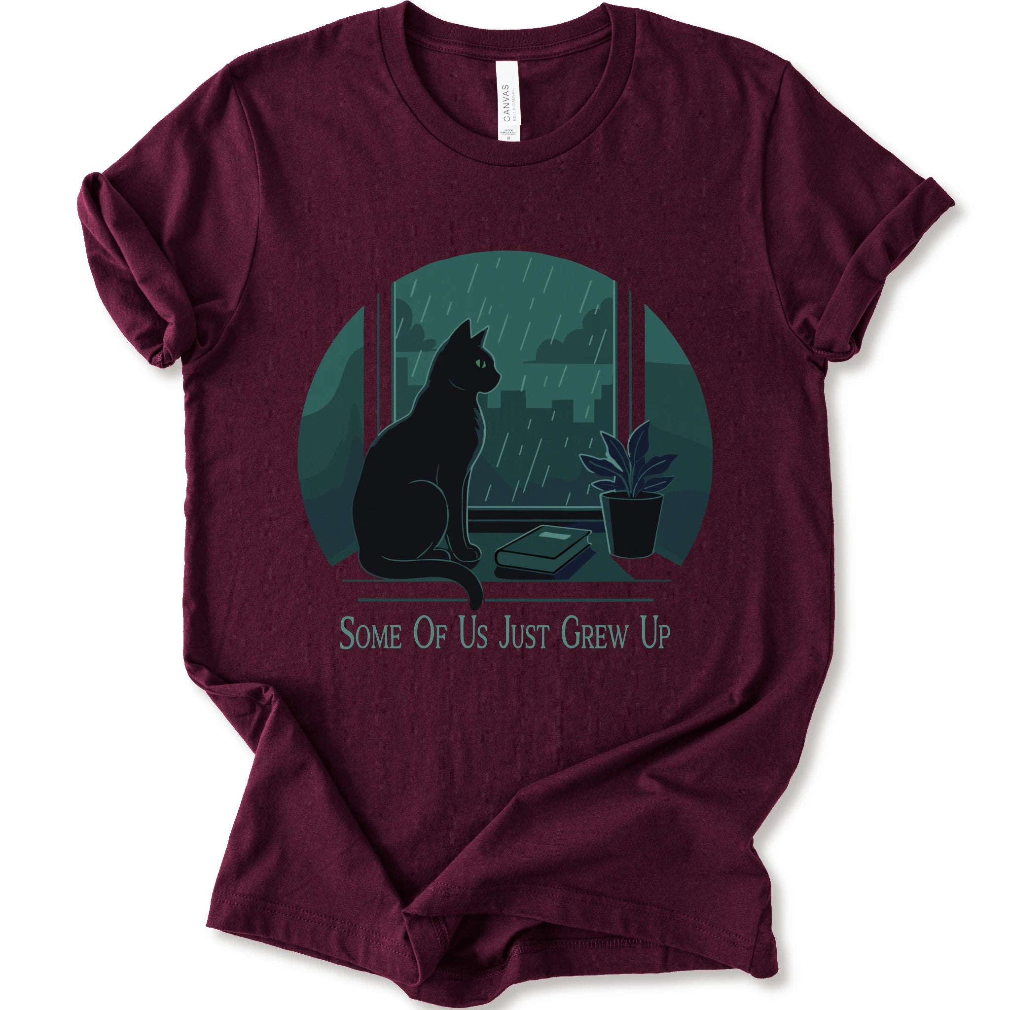 A maroon short-sleeve crewneck t-shirt with a graphic print of a black cat sitting by a window in the rain, next to a small potted plant, with the text 'SOME OF US JUST GREW UP' printed below.